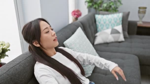 落ち着いたアジアの女性は静けさと快適さを描いたスタイリッシュなリビングルームでモダンなソファーで平和な瞬間を楽しんでいます — ストック動画