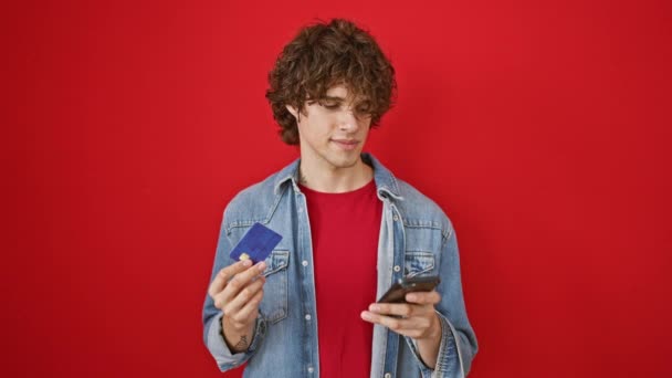 有一头卷发的英俊年轻人拿着信用卡和红色背景的智能手机 — 图库视频影像