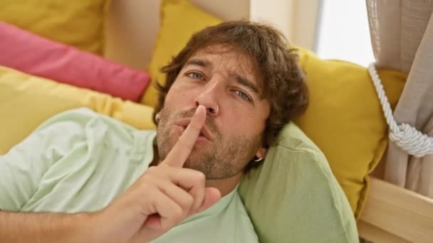 秘密在年轻 迷人的高加索男人穿着睡衣躺在家里讲故事时 用手指捂住嘴唇做着 的手势 卧室的故事时间 — 图库视频影像
