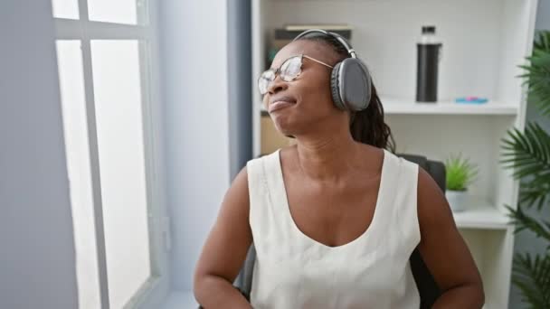 头发卷曲的非洲裔美国妇女 在明亮的办公室室内欣赏着耳机的音乐 — 图库视频影像