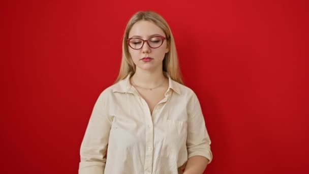 神经质的年轻金发女郎戴着眼镜为问题烦恼 皱着眉头表示怀疑 在红色背景下被隔离的消极人士 — 图库视频影像