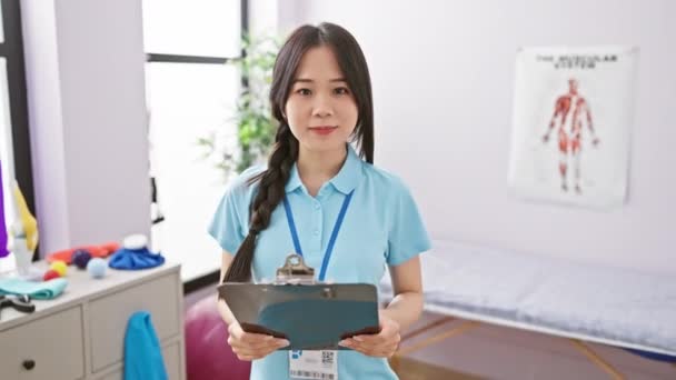 Smiling Asian Woman Blue Healthcare Uniform Holding Clipboard Rehabilitation Clinic — Stock Video