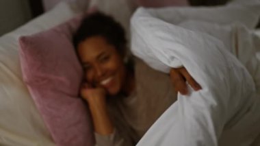 Kıvırcık saçlı, pembe yastıklarla ve beyaz çarşaflarla çevrili, rahat bir yatak odasında rahat bir şekilde yuvalanmış gülümseyen bir kadın..