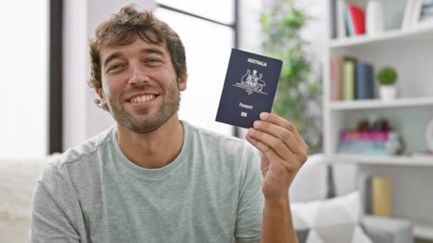陽気で自信に満ちた若い男性は オーストラリアのパスポートを誇らしげに保持し 喜んで笑顔で彼のリビングルームソファに座って — ストック動画