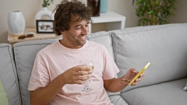 用智能手机微笑的男人 把酒杯放在舒适的客厅里 — 图库视频影像