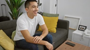 Gülümseyen İspanyol genç adam, modern bir oturma odasında, elinde uzaktan kumandayla rahatça oturuyor.