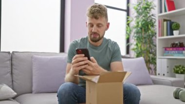 Beyaz sakallı genç bir adam gri kanepeli rahat bir oturma odasında akıllı telefon kullanırken gülümsüyor..