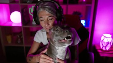Gülümseyen orta yaşlı, gri saçlı kadın flaması, sallanan kulaklıklar ve köpeği canlı bir oyun odasında, dijital eğlencenin tadını çıkarıyor.