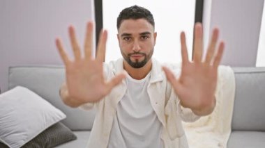 Neşeli genç Arap adam parmaklarıyla on numarayı gösteriyor. Evinin kanepesinde rahatça otururken mutlu bir şekilde işaret ediyor, pozitif titreşimler yayıyor.