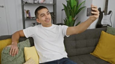 Çekici İspanyol adam rahat bir oturma odasında gri bir kanepede dinlenirken selfie çekiyor..