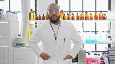 Laboratuvar önlüklü sakallı, kendine güveni tam bir adam. Parlak bir laboratuarda duruyor, kolları çapraz, güvenlik gözlükleriyle..