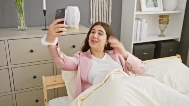 Gülümseyen kadın yatak odasında akıllı telefonuyla selfie çekiyor günlük yaşam tarzı ve modern ev dekorasyonu sergiliyor..