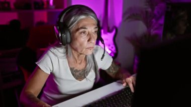 Stresli orta yaşlı, gri saçlı kadın flamalı evinde, karanlık bir odada bilgisayarında oyun oynamanın stresiyle savaşıyor.