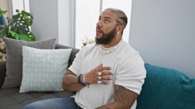Gururlu Afro-Amerikan adam oturma odasındaki kanepede oturuyor, dostane bir gülümseme ve göğsünde ellerle. Minnettarlığın ve evde sağlığın kapalı bir portresi.