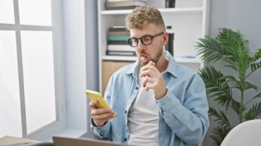 Gözlüklü genç sakallı bir adam modern ofis ortamında yemek yerken akıllı telefon kullanır..