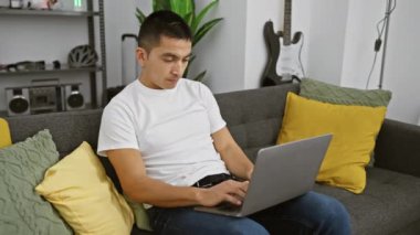 Genç İspanyol adam, modern oturma odasındaki koltukta dizüstü bilgisayar kullanıyor. Konfor ve bağlantı gösteriyor..