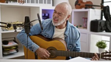 Modern ev stüdyosunda gitar çalıp şarkı söyleyen olgun sakallı bir adam.