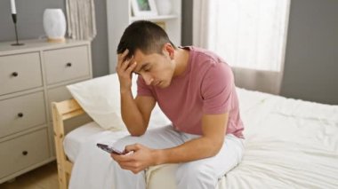 Pembe tişörtlü genç bir İspanyol iyi ışıklandırılmış bir yatak odasında yatağında telefonuna bakarken sıkıntılı görünüyor..