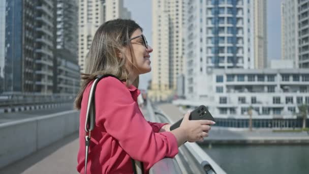 戴着眼镜 穿着粉色衬衫 拿着手机 背对着杜拜码头摩天大楼的微笑的女人 — 图库视频影像