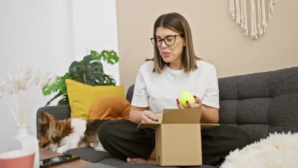 一位快乐的年轻女子在舒适的客厅里打开一只盒子 旁边躺着一只狗 — 图库视频影像
