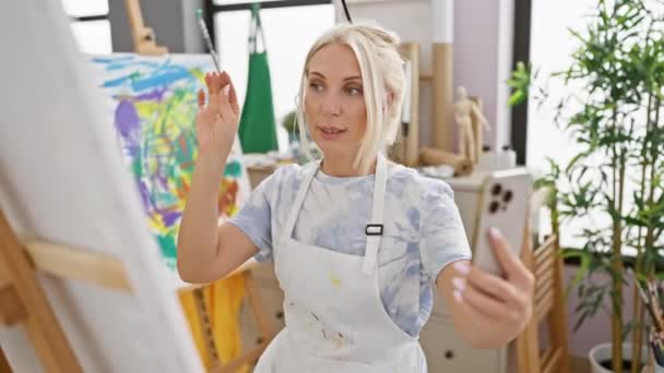 年轻而自信的金发女画家在一个视频通话中描绘着自己 全神贯注地在艺术工作室的室内上着快乐的课 巧妙地挥动画笔和调色板 — 图库视频影像