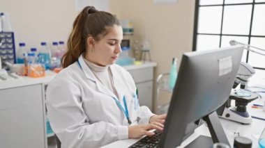 Odaklanmış bir İspanyol kadın bilim adamı bir laboratuvarda bilgisayar başında çalışıyor, profesyonellik ve kararlılık gösteriyor..