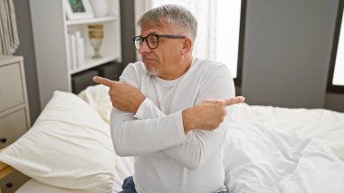 Gri saçlı bir adam, aydınlık bir yatak odasında bir yatakta oturuyor, kafası karışmış ve iki farklı yöne bakıyor..