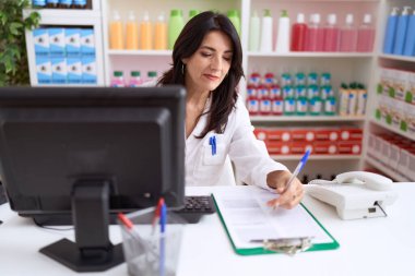 Middle age hispanic woman pharmacist writing on document using computer at pharmacy