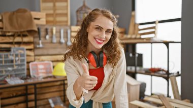 Gülümseyen genç marangoz, kereste zengini marangozluk atölyesinde baş parmak hareketi yapıyor.
