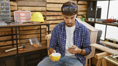 Yakışıklı genç bir Arap adam, mesleği marangoz, canlı marangozluk atölyesinde akıllı telefonuyla mesajlaşırken kahvesinin tadını çıkarıyor.