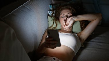 Genç bir kadın geceleri akıllı telefonunu kullanarak yatağında uzanıyor, modern kapalı mekan yaşam tarzı ve teknoloji kullanımını resmediyor..