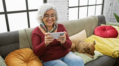 Gülümseyen gri saçlı bir kadın rahat bir oturma odasında dinlenen köpeğinin yanındaki koltukta otururken akıllı telefon kullanmaktan zevk alır..
