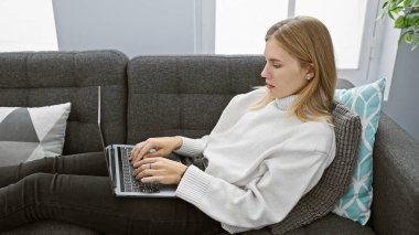 Odaklanmış sarışın bir kadın, iyi ışıklandırılmış bir evin içindeki gri bir koltukta otururken rastgele dizüstü bilgisayar kullanıyor..