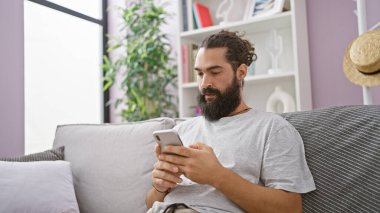 Modern oturma odasındaki rahat koltukta akıllı telefon kullanan sakallı yakışıklı İspanyol bir adam.