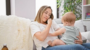 Anne akıllı telefonda konuşuyor, evde kızıyla koltukta oturuyor.