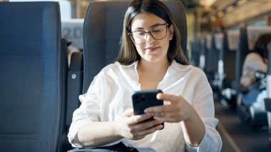 Genç, güzel, İspanyol bir kadın gülümsüyor. Akıllı telefon kullanıyor. Trende oturuyor.