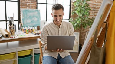 Gülümseyen İspanyol bir adam yaratıcı bir sanat stüdyosunda dizüstü bilgisayar kullanıyor ve etrafta resimler ve malzemeler var.