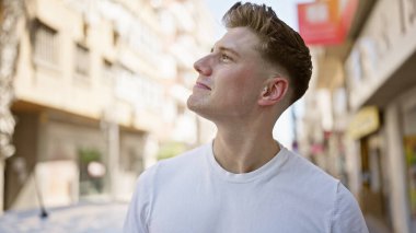 Yakışıklı beyaz bir adam, canlı bir şehir caddesinde duruyor, yukarı bakıyor, kendine güvenerek gülümsüyor, gökyüzüne neşe saçıyor, havalı, pozitif bir yaşam tarzını yansıtıyor..