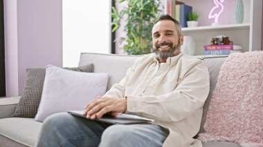 Oturma odasındaki rahat bir kanepede elinde tabletle gülümseyen olgun sakallı adam..