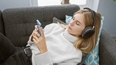 Rahat, mavi gözlü, kulaklık takan sarışın bir kadın rahat bir oturma odasında akıllı telefonuyla müzik dinlemekten hoşlanıyor..