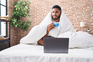 Young latin man using laptop drinking coffee at bedroom