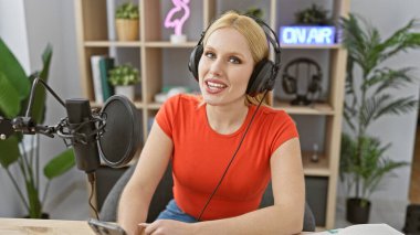 Kırmızılı sarışın kadın radyo stüdyosunda mikrofona konuşurken gülümsüyor.
