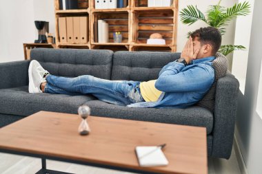 Young hispanic man patient stressed lying on sofa at psychology clinic
