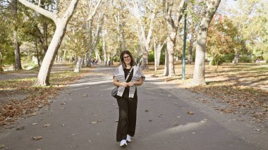 Güneşli bir sonbahar parkında, ağaçlar ve yapraklarla çevrili gülümseyen genç İspanyol kadın..