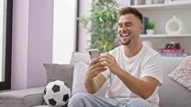 Evde futbol topuyla kanepede akıllı telefon kullanan gülümseyen adam.