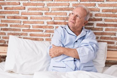 Orta yaşlı, kır saçlı, mutsuz bir ifadeyle yatakta oturan ve yatak odasında el kol hareketleri yapan bir adam.