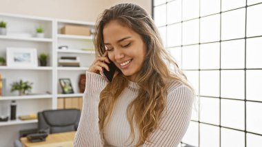 Kendinden emin ve mutlu İspanyol bir kadın, başarılı bir iş adamı, akıllı telefonuyla sohbetin tadını çıkarırken zarif ofis içinde, iş yerinde pozitiflik yayıyor.