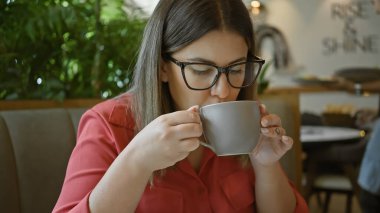 Genç, esmer bir kadın, gözlüklü, parlak bir kafede kahvesini yudumluyor..