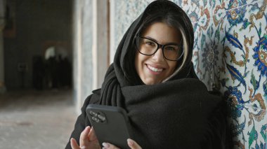 İstanbul 'da gözlük ve başörtüsü takan gülümseyen genç bir kadın süslü fayanslarla akıllı telefon kullanıyor..