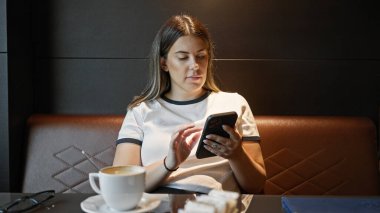 Genç bir esmer kadın kapalı bir kafe masasında tek başına kahve içerken akıllı telefon kullanıyor..